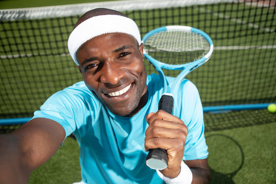 Top view portrait of smiling man taking selfie with camera. He is sitting on court and holding racket. Guy is relaxing after match