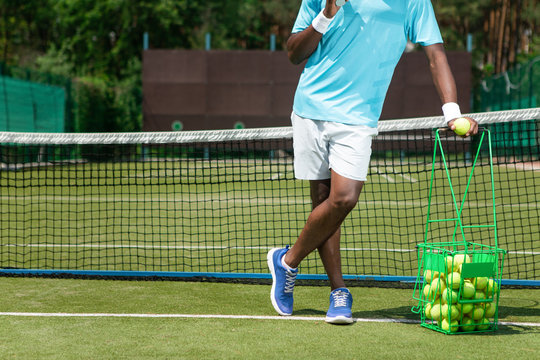 Shapely man is standing near net with basket of balls. He is spending active time playing sport. Copy space in left side