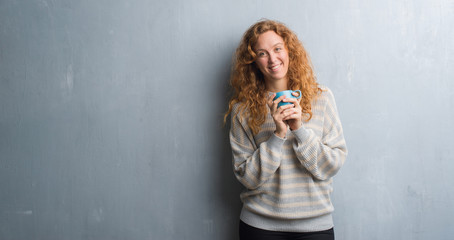 Young redhead woman over grey grunge wall drinking a cup of coffee with a happy face standing and smiling with a confident smile showing teeth