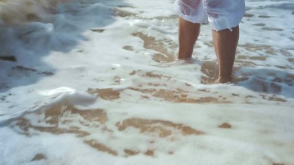 Sea Wave Washes the Elder Woman's Legs in White Trousers