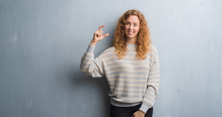 Fototapeta premium Young redhead woman over grey grunge wall smiling and confident gesturing with hand doing size sign with fingers while looking and the camera. Measure concept.