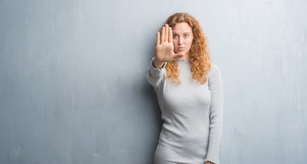 Fototapeta premium Young redhead woman over grey grunge wall with open hand doing stop sign with serious and confident expression, defense gesture
