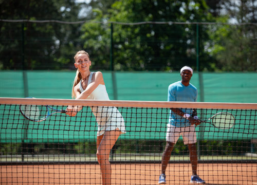 Focus on cheerful woman receiving ball with racket during tennis match. She is enjoying game with male partner who is standing behind. Friends are doing sport in one team