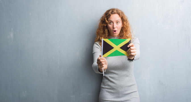 Young redhead woman over grey grunge wall holding flag of Jamaica scared in shock with a surprise face, afraid and excited with fear expression