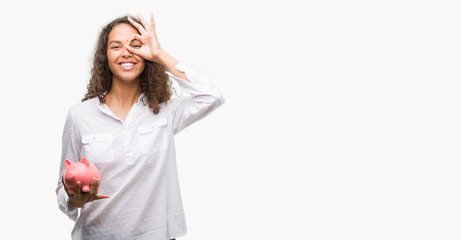 Young hispanic woman holding piggy bank with happy face smiling doing ok sign with hand on eye looking through fingers