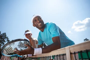 Poster Low angle of delighted man relaxing during break in double tennis set. He is standing with his female co-player and they are leaning against net. Athletes are keeping rackets while looking forward to © Yakobchuk Olena
