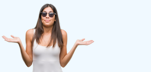 Young beautiful hispanic wearing sunglasses clueless and confused expression with arms and hands...