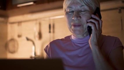 Senior woman working on laptop from home office in evening. Smiling retired female freelancer using mobile phone to discuss project with colleague at night. Overworked businesswoman in kitchen at home