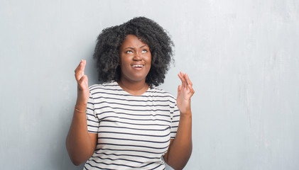 Young african american plus size woman over grey grunge wall crazy and mad shouting and yelling with aggressive expression and arms raised. Frustration concept.