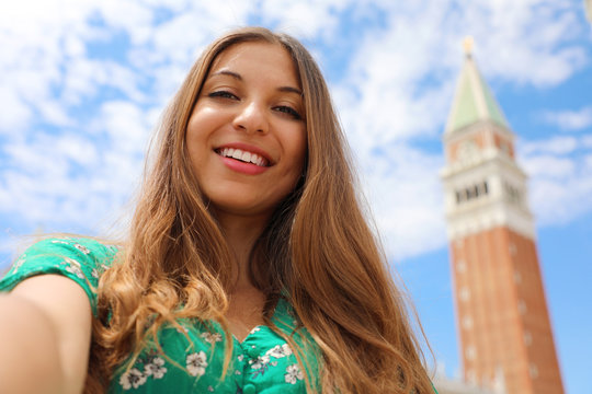 Happy beautiful woman taking selfie photo in Venice with white clouds in the sky. Tourist girl smiling at camera in St. Mark square with bell tower in Venice, Italy.