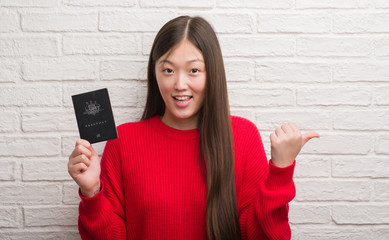 Young Chinese woman over brick wall holding passport of Australia pointing and showing with thumb up to the side with happy face smiling