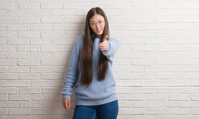 Young Chinise woman over white brick wall smiling friendly offering handshake as greeting and welcoming. Successful business.