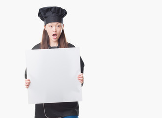Young Chinese woman over isolated background wearing chef uniform holding banner scared in shock with a surprise face, afraid and excited with fear expression