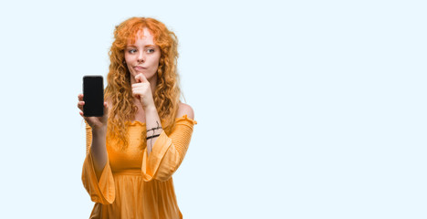 Young redhead woman showing smartphone serious face thinking about question, very confused idea