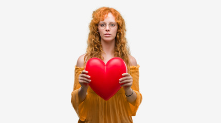 Young redhead woman in love holding red heart with a confident expression on smart face thinking serious