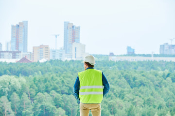 Rear view of male construction engineer in white hardhat and green vest contemplating forests and...