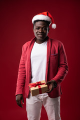 Favorite holiday. Smiling guy in santa hat posing with new year present on red background