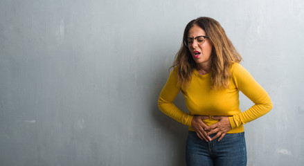 Fototapeta premium Middle age hispanic woman over grey wall wearing glasses with hand on stomach because indigestion, painful illness feeling unwell. Ache concept.