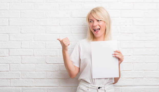 Adult caucasian woman over white brick wall holding blank paper sheet pointing and showing with thumb up to the side with happy face smiling