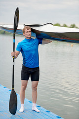 Athletic young man standing with sport boat and paddle on the pier outdoors