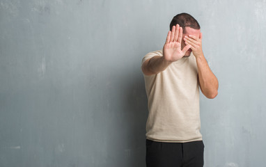Young caucasian man over grey grunge wall covering eyes with hands and doing stop gesture with sad and fear expression. Embarrassed and negative concept.