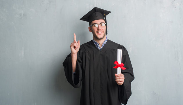 Young caucasian man over grey grunge wall wearing graduate uniform holding degree surprised with an idea or question pointing finger with happy face, number one