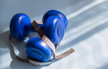 Blue dumbbells, tape measure on a blue background in the sun. Healthy lifestyle, the concept of losing body weight. Cares about the body. Empty place for text.