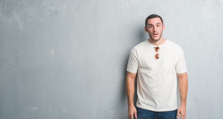 Young caucasian man over grey grunge wall wearing sunglasses afraid and shocked with surprise expression, fear and excited face.