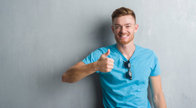 Young redhead man over grey grunge wall wearing casual outfit doing happy thumbs up gesture with hand. Approving expression looking at the camera with showing success.
