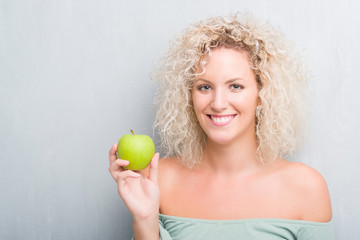 Young blonde woman over grunge grey background eating green apple with a happy face standing and smiling with a confident smile showing teeth