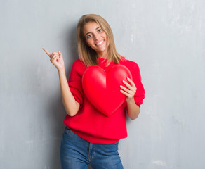 Beautiful young woman over grunge grey wall holding red heart very happy pointing with hand and finger to the side