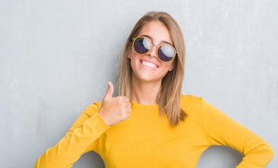 Beautiful young woman standing over grunge grey wall wearing retro sunglasses happy with big smile...
