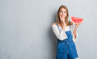 Beautiful young woman over grunge grey wall eating water melon serious face thinking about...