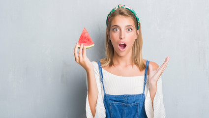 Beautiful young woman over grunge grey wall eating water melon scared in shock with a surprise...