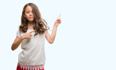 Brunette hispanic girl smiling and looking at the camera pointing with two hands and fingers to the side.