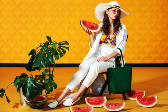 Full Body Studio Fashion Portrait Of Young Beautiful Model Wearing Long White Trench Coat, Trousers, Wide-brimmed Hat, Loafers, Yellow Sunglasses, Holding Green Bag, Posing On  Background. Copy Space