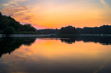 Golden sunset on a pond. Mysterious moment when the orange sun says goodbye to nature. Its last reflection trembles on the water. Evening twilight becomes darker more and more.