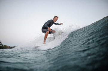 Young wakeboarder riding down the blue wave against the background of clear sky