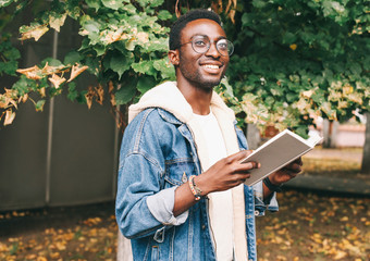 Fototapeta premium Happy young smiling african man holds book in autumn city park
