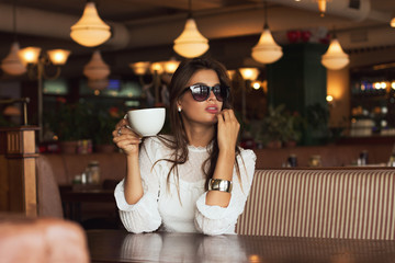Beautiful girl in glasses is drinking coffee at table in cafe. Emotional portrait, film effect, author's processing.