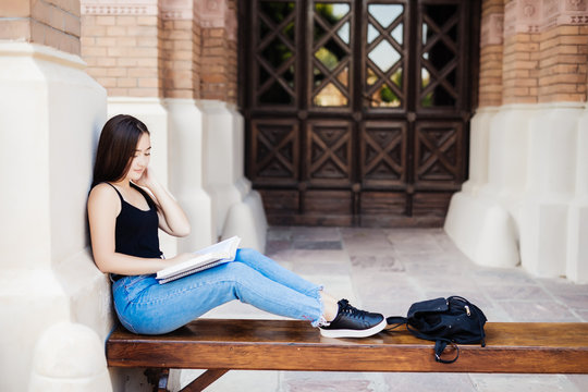 Smiling Young Asian Woman Sitting On A Bench Studing In The Park. Brunette Asian Girl Relaxing And Reading A Book. Lifestyle And Chiling Concept