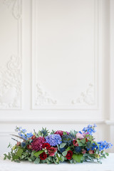 Elongated floral arrangement in vintage metal vase. table setting. Blue and red color. Gorgeous bouquet of different flowers. Bright room, stucco on the wall