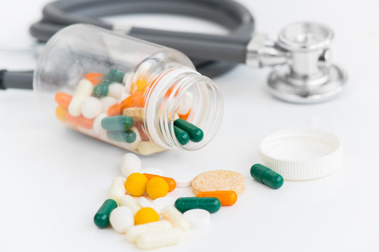 Close up of assorted colorful medication drugs and vitamins spilling from bottle over white background next to stethoscope in doctors office