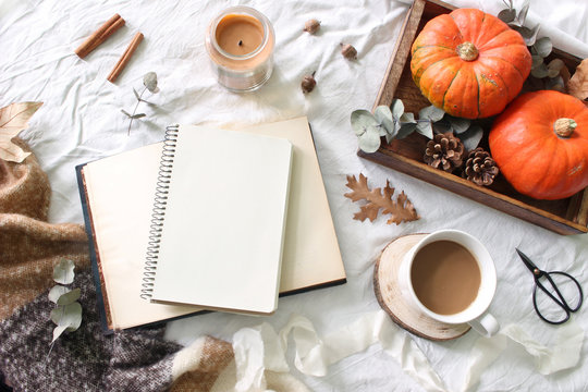 Autumn breakfast in bed composition. Blank notepad, book mockup. Coffee, candle,eucalyptus leaves and pumpkins on wooden tray. White linen bed sheet background. Thanksgiving, halloween. Flat lay, top.