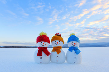 Three small snowman in colorful hats are standing on the snow. The scenery with mountains, field in...