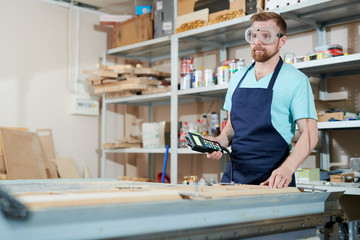 Artisan in glasses and apron using machine while working with wood in carpenters shop