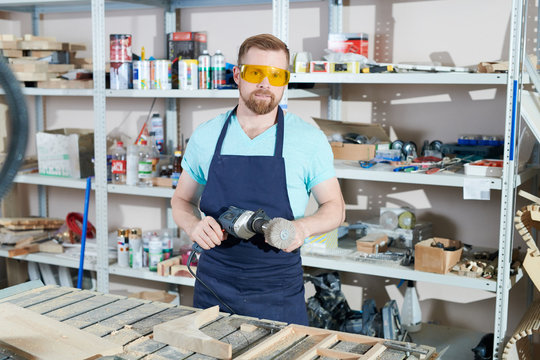 Portrait of bearded young carpenter wearing safety goggles and apron standing with electric drill in workshop