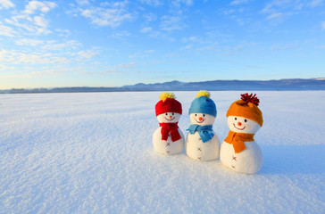 Three small snowman in colorful hats are standing on the snow. The scenery with mountains, field in...