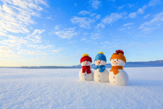 Friends snowman in red, blue, yellow hats and scarfs. Nice winter scenery with mountains, field in snow. Orange sunset background.