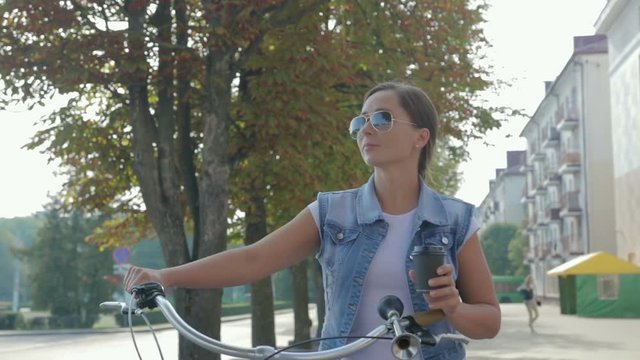Image of a beautiful, young girl with a pigtail, in a denim suit and sunglasses, who walks leisurely down the street with a glass of coffee and rolls a purple Bicycle.